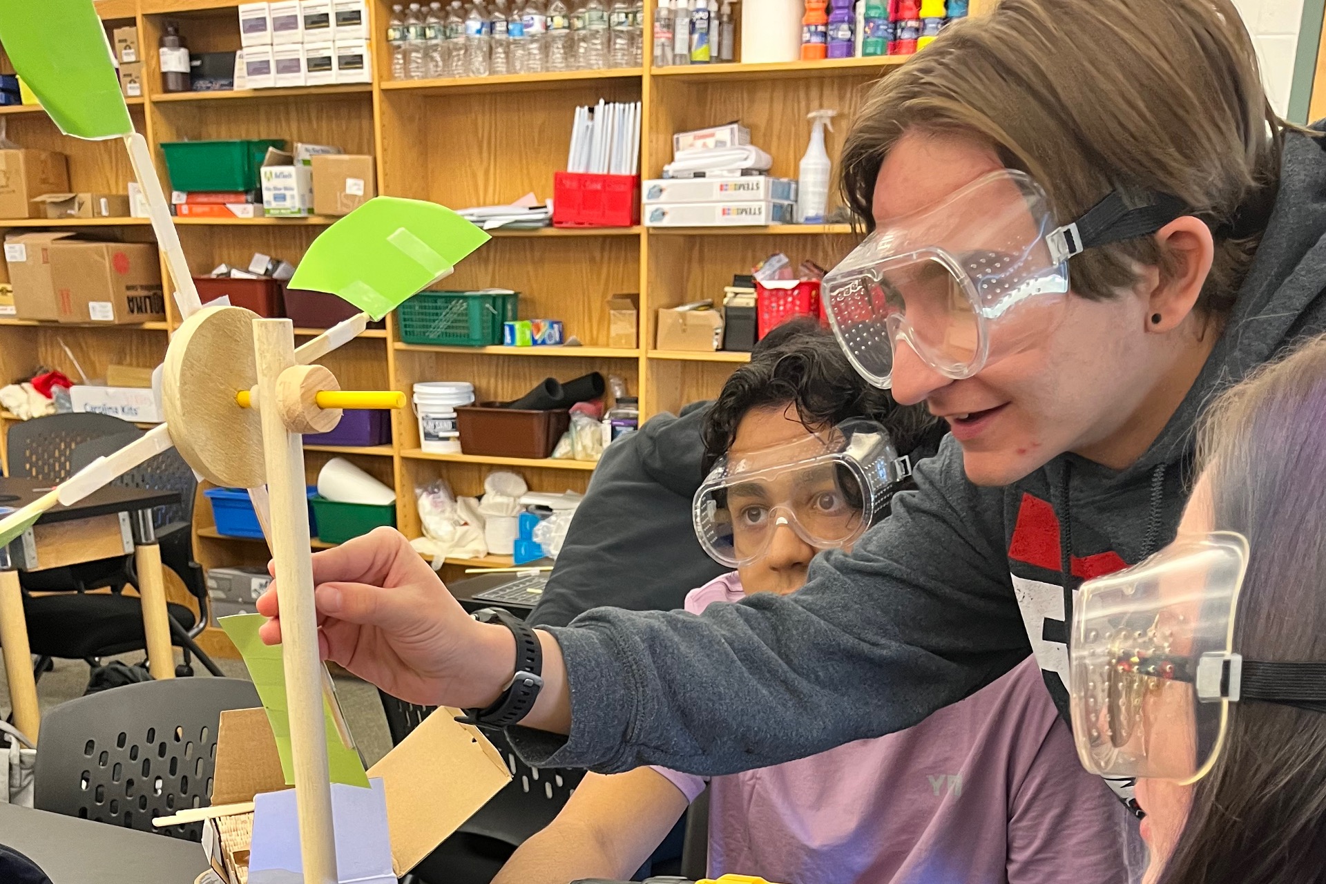 Teacher and students looking at wind turbine model built by wood pieces and paper in a class room.